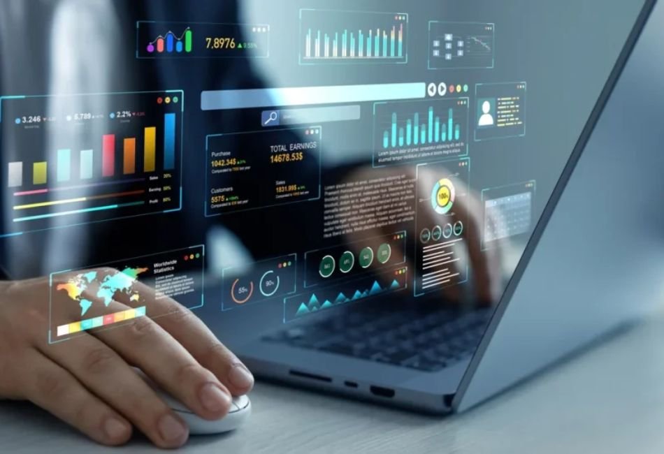 A professional working on a laptop, using a mouse while an interactive digital overlay displays futuristic business analytics, financial dashboards, bar charts, line graphs, and world map statistics over the screen.